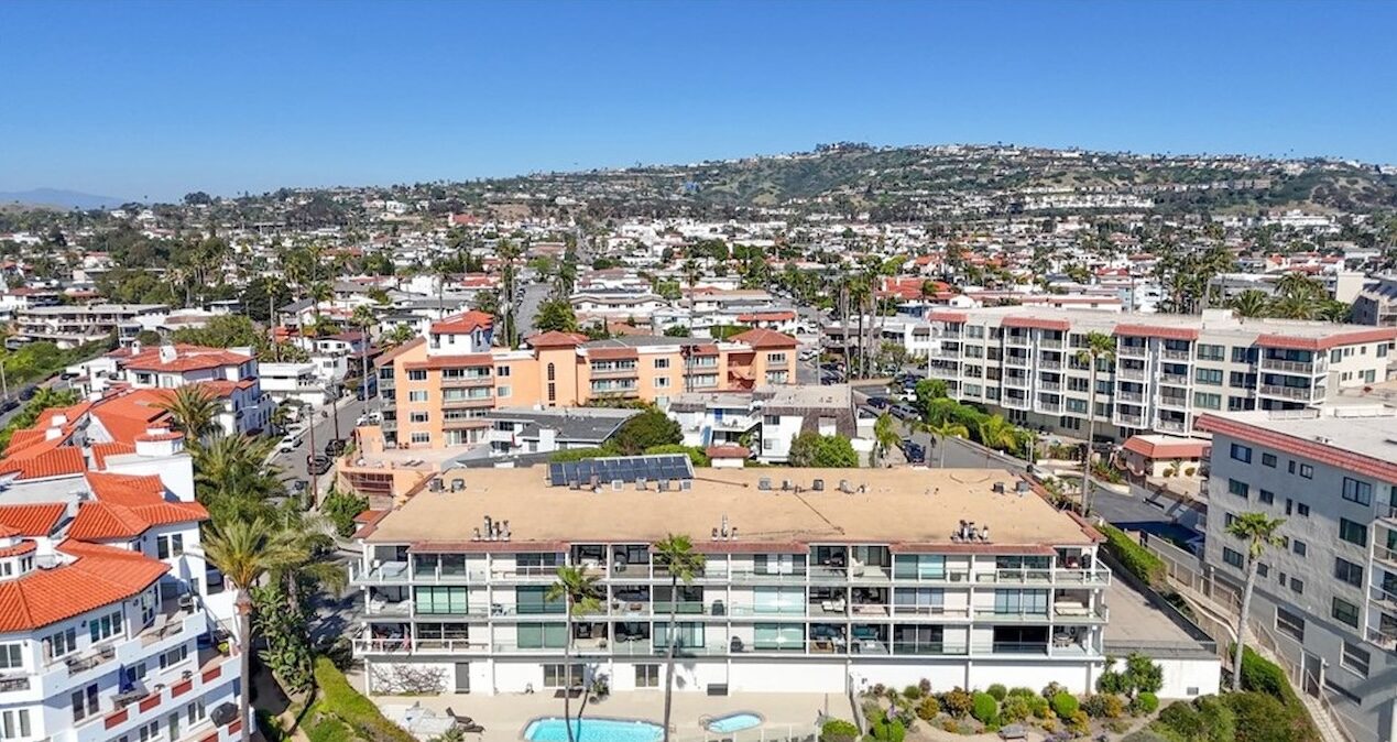 Aerial view of Spanish-style coastal buildings with terracotta roofs, palm trees and pools, with hills behind in San Clemente