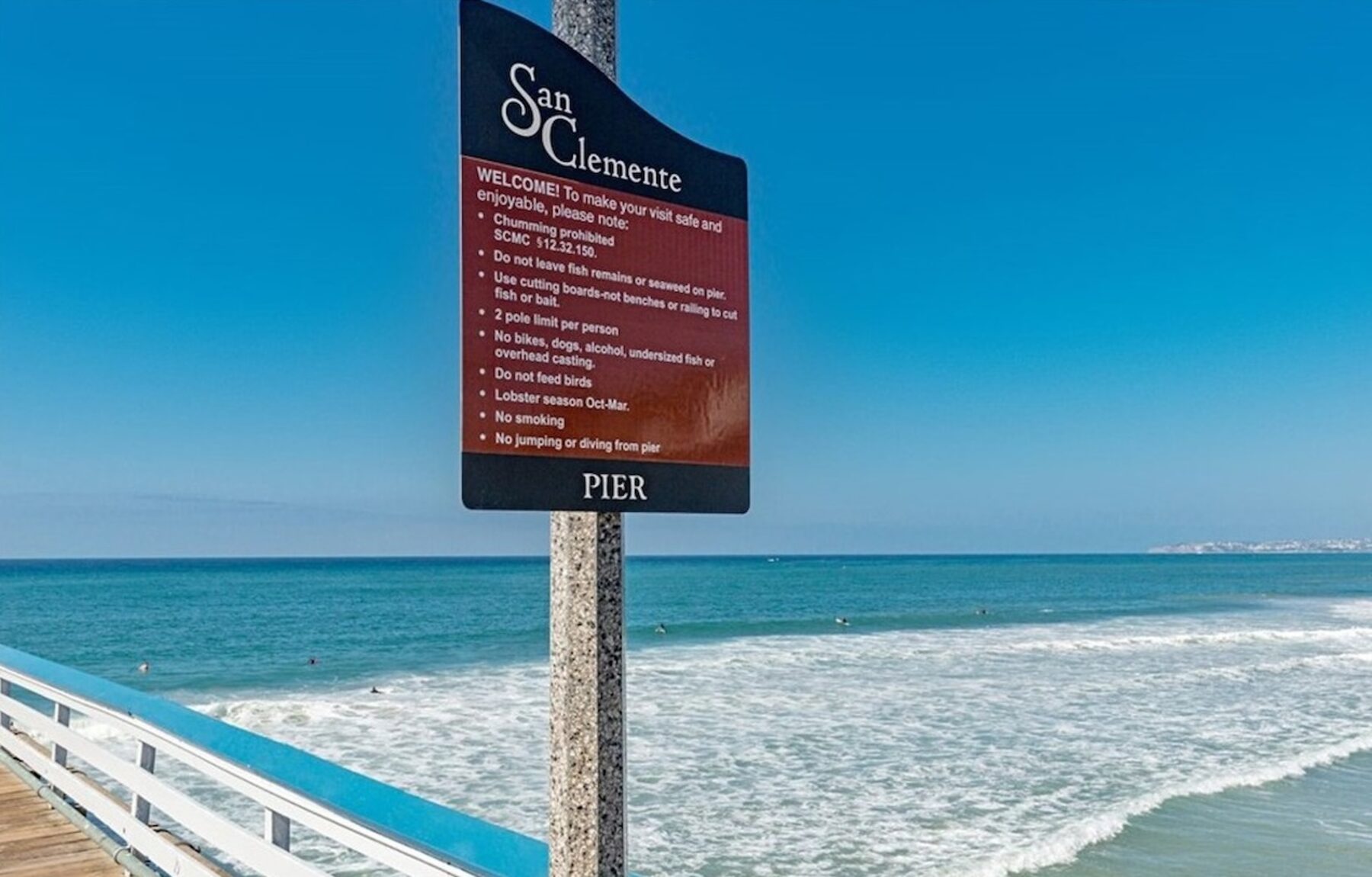 San Clemente pier welcome sign beside a railing, looking out over blue ocean with surfers in the water and white wash rolling in