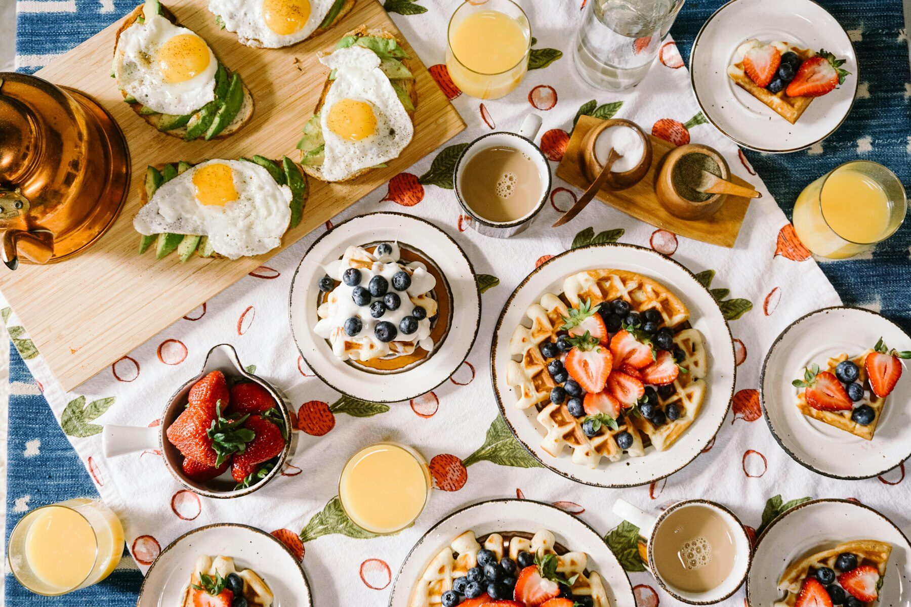 Brunch table spread with waffles topped with berries, avocado toast with fried eggs, orange juice and coffee